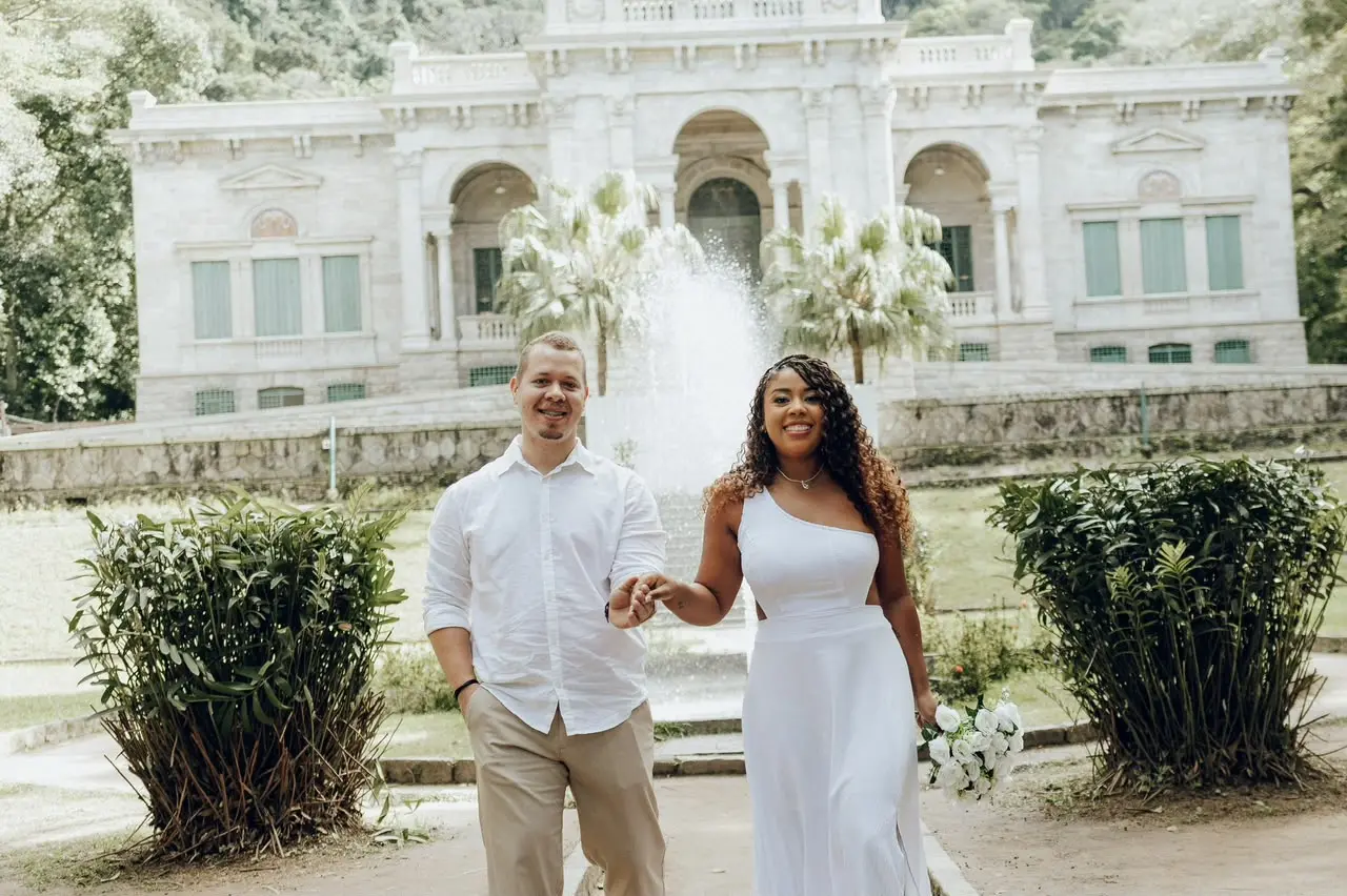 Mayara e Caio de mãos dadas, de branco, em frente a um palacete com chafariz